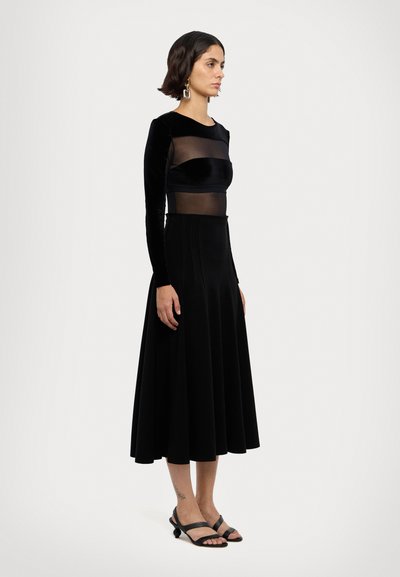 Robe en velours noir avec des manches longues et un corsage transparent. Jupe évasée avec des plis ; portée avec des sandales noires à brides grises.