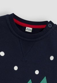 Jūras zilas džemperis ar baltām izšūtām punktiņām, zaļš trīsstūra dizains, sarkans iekšējais apkakle un pogu detaļa uz pleca.