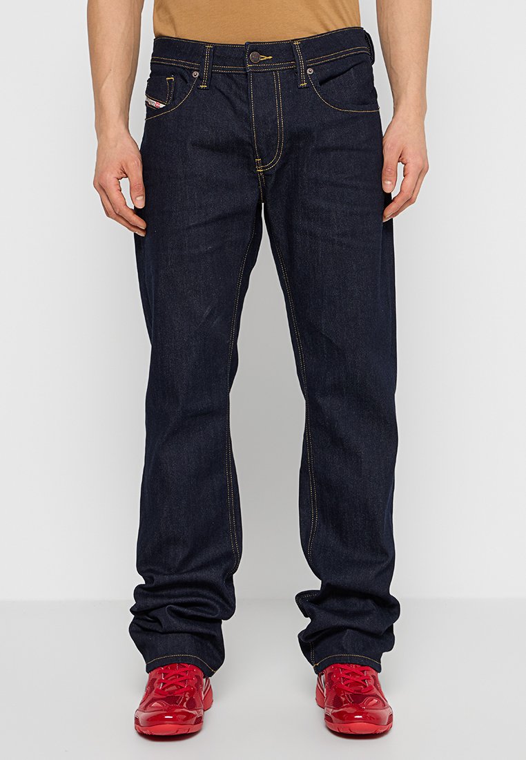 Dunkelblaue Denim-Jeans mit gelben Kontrastnähten, Fünf-Taschen-Design und Knopfverschluss. Getragen mit roten Turnschuhen.