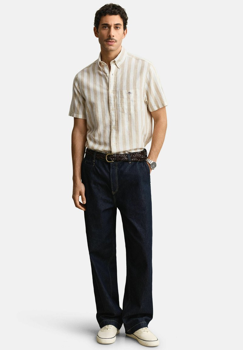 Mann trägt beige-weiß gestreiftes kurzärmeliges Hemd, dunkelblaue Jeans mit geflochtenem braunem Gürtel, weiße Turnschuhe und silberne Armbanduhr.