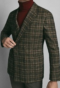 Blazer a quadri marrone con una vestibilità strutturata, rever a punta, due bottoni e un tessuto intrecciato testurizzato, indossato sopra un dolcevita rosso scuro.
