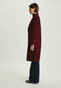 Manteau long bordeaux avec motif floral texturé, manches larges et devant ouvert. Porté sur un pull à col roulé noir et un pantalon en denim évasé.