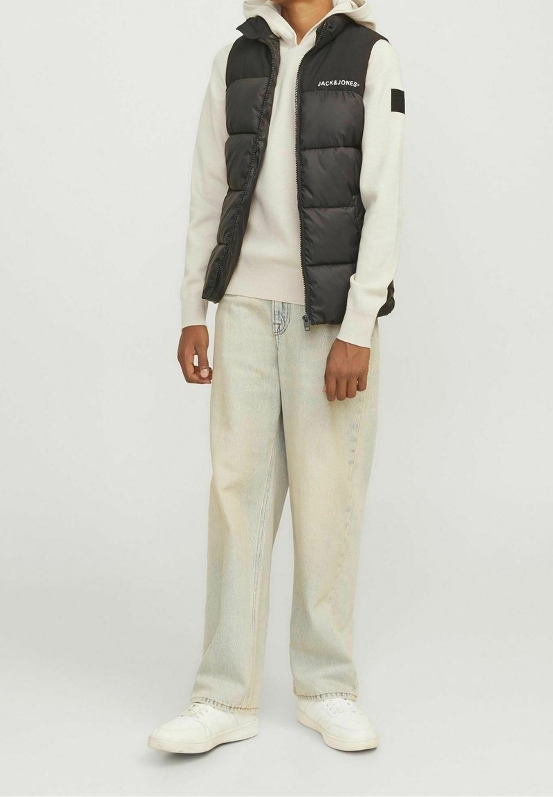 Personne portant un gilet matelassé noir sur un sweat à capuche beige avec un jean ample délavé clair et des baskets blanches sur un fond uni.