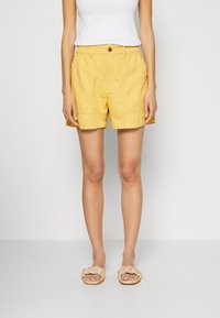 Kvinna som bär ljusgula shorts med framfickor, vit ärmlös topp och beige flätade sandals, står framför en ensam bakgrund.