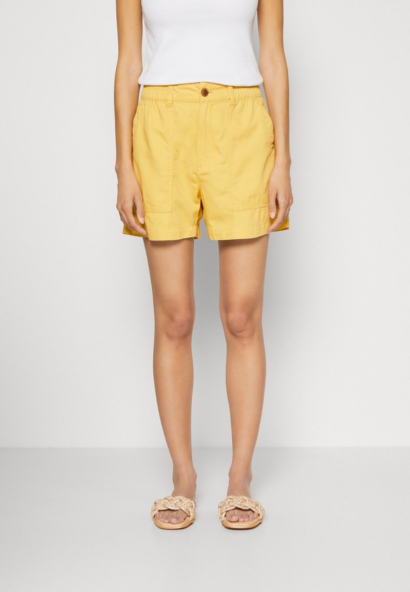 Kvinna som bär ljusgula shorts med framfickor, vit ärmlös topp och beige flätade sandals, står framför en ensam bakgrund.