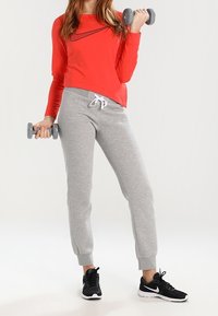 Roter langärmeliger Sporttop mit schwarzem Akzent, kombiniert mit grauen Jogginghosen und schwarzen Sneakern. Das Model hält graue Hanteln.