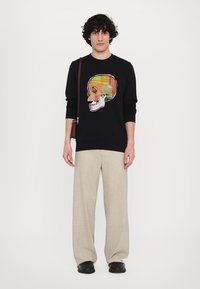 Sort sweatshirt med en farverig kraniegrafik, parret med lyse beige bukser med bred ben og sorte sko. Modellen bærer en mørk skuldertaske.