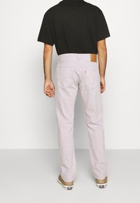 Hombre vestido con jeans rectos de color lavanda claro, una camiseta negra de manga corta y zapatillas beige, de pie frente a un fondo liso.