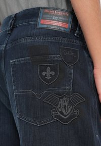 Σκούρα τζιν από ντένιμ με ξεβάμματα. Διαθέτουν κεντημένα εμβλήματα: ένα σήμα ασπίδας, ένα fleur-de-lis και έναν αετό στη πίσω τσέπη.