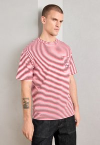 Rød og hvit stripet t-skjorte med korte ermer, rund hals og en brystlomme med brodert tekst. Dongeribukser er synlige.