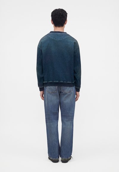 Homme debout de dos, portant une veste en jean bleu foncé et un jean bleu ample sur un fond blanc uni.