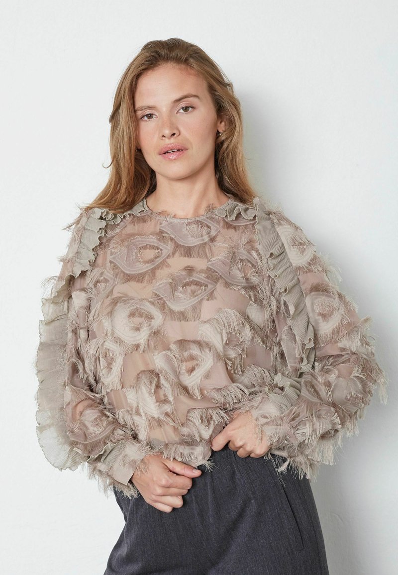 Lehká průsvitná blůza v béžové barvě s texturovaným květinovým vzorem, zdobená volánkovými okraji a dlouhými rukávy. V kombinaci s tmavými elegantními kalhotami.