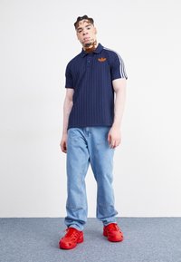 Man med vitiligo som bär en marinblå Adidas-polo, ljusblå jeans och ljusröda sneakers står mot en vit vägg och en grå heltäckningsmatta.