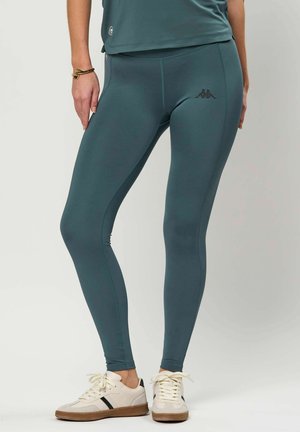 Femeie purtând colanți verde-albăstrui și adidași albi, stând cu o mână sprijinită pe coapsă, pe un fundal simplu.