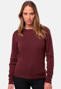 Femme aux cheveux raides châtains clairs portant un pull à manches longues bordeaux uni et un pantalon noir, se tenant devant un fond blanc.