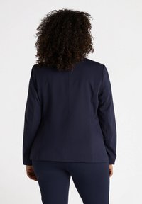 Laivastonsininen bleiseri, jossa on räätälöity istuvuus, rakenteelliset hartiat, pitkät hihat ja sileä kangas. Yksinkertainen muotoilu ilman näkyviä kuvioita tai koristeita.