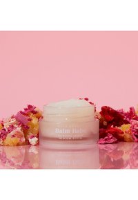 Genomskinlig glasburk med läppbalsam med vit krämig konsistens, omgiven av färgglada smulor på en rosa bakgrund. Förpackningen är märkt "Balm Babe."