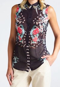 Blusa sin mangas negra con estampado floral en rojo y verde, cuello con tachuelas, cierre de botones en la parte delantera y dos bolsillos frontales.