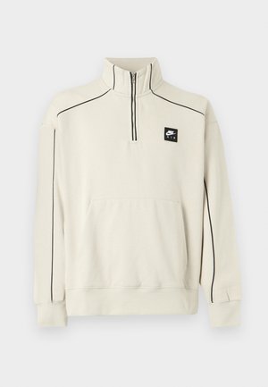 Sweat-shirt beige clair avec un col demi-zip, des coutures décoratives noires et une poche avant, arborant un logo Nike Air noir sur la poitrine.