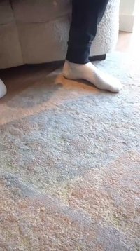 Calcetín blanco en un pie desnudo, apoyado sobre una alfombra de color claro con textura y patrones sutiles. Pantalones oscuros visibles por encima del tobillo.