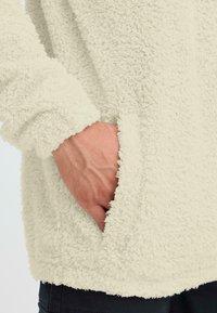 Flauschiger beiger Pullover mit langen Ärmeln, der eine Seiten Tasche und strukturierter Stoff aufweist, und eine weiche, plüschige Oberfläche betont.
