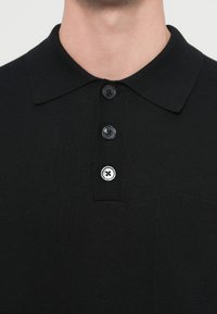 Czarna dzianinowa koszulka polo z klasycznym kołnierzykiem, trzema czarnymi guzikami i małą przednią kieszonką. Tkanina jest fakturowana i lekka.