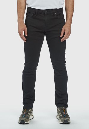 Mees, kes kannab kitsaid musti teksaseid ja must-beez sneakers'e, seisab lihtsalt heleda tausta ees.