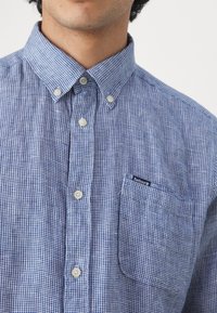 Homme portant une chemise à carreaux bleu et blanc avec une poche poitrine portant l'étiquette Barbour, visible du cou jusqu'au milieu du torse.