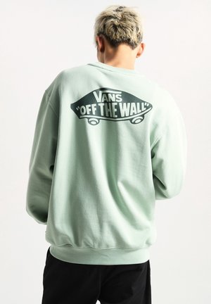 Osoba z krótkimi blond włosami nosi miętową bluzę Vans z czarnym logo deskorolki "Off The Wall" na plecach, zwrócona tyłem.