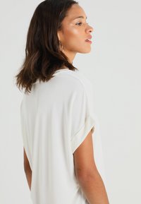 Laisvai krentantis baltas marškinėlių viršus iš lengvos medžiagos, su suvyniotomis rankovėmis ir apvaliu iškirpimu. Lygus paviršius su minimaliais dizaino akcentais.