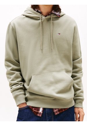 Osoba ubrana w jasnooliwkową bluzę z kapturem z przednią kieszenią, pod którą ma czerwoną, kraciastą koszulę oraz niebieskie dżinsy, z rękami częściowo w kieszeniach.