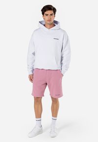 Hvid hættetrøje med forlommer og sort logo, parret med lyserøde shorts med sidelommer og logo. Hvide sneakers og sokker.