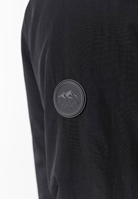 Tela negra con un acabado texturizado que presenta un parche de goma redondo. El parche muestra montañas y el texto "CLOSURE LDN, OUTDOOR GEAR, EST 2015".