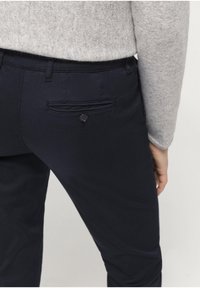 Mörkblå byxor med en bakficka med knapp, tillverkade av slät tyg. Midjan är platt och passformen är rak.