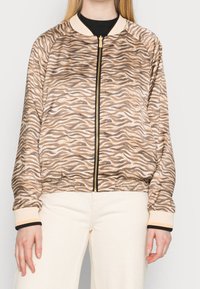 Beige bomberjakke med sorte zebra-striber, ribbede manchetter og en guldfarvet lynlås. Letvægtsmateriale med elastisk talje, der har en afslappet pasform.