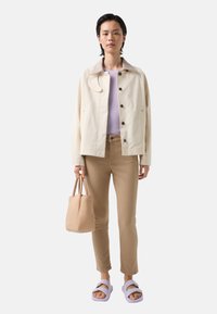 Light beige jacket with a collared neck, black buttons, lavender top, beige pants, light purple sandals, and a small tan handbag.