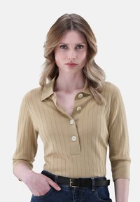 Khakifarbener Rippenstrick-Pullover mit kragenförmigem Halsauschnitt und drei Knöpfen. Hat halblange Ärmel und wird mit dunklen Jeans und einem schwarzen Gürtel kombiniert.