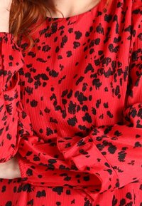 Rote plissierte Bluse mit schwarzem Leopardenmuster. Lange Ärmel mit gerafften Bündchen und Rundhalsausschnitt. Subtile glänzende Textur.