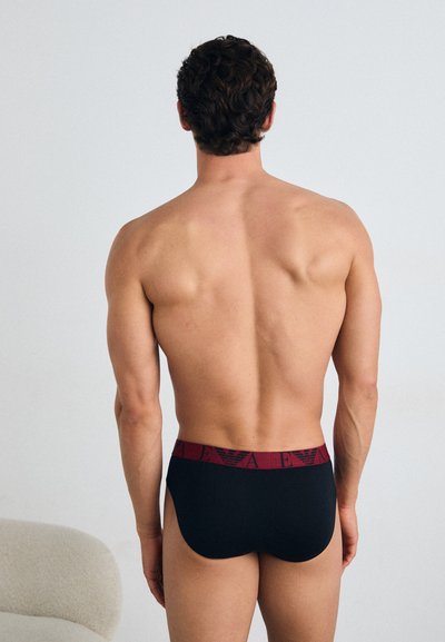 Schwarze Boxer Shorts mit einem burgunderfarbenen elastischen Bund, der ein Logo trägt. Der Stoff wirkt glatt mit einer engen Passform und wird von hinten gezeigt.