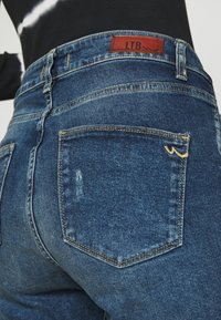 Jeans de mezclilla azul oscuro con cintura alta, que presentan un parche de cuero marrón claro etiquetado "LTB" y costuras amarillas en el bolsillo trasero.