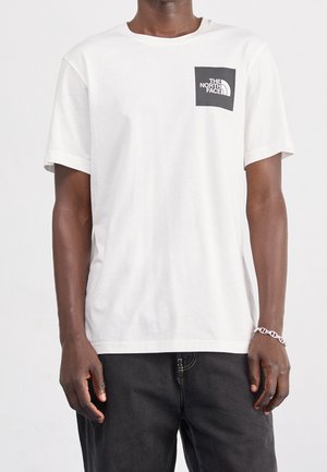 Mand iført en hvid t-shirt med korte ærmer og et sort firkantet North Face-logo på brystet, kombineret med mørke bukser og et sølvarmbånd.