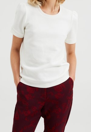 Nainen pukeutuneena valkoiseen, teksturoituun lyhythihaiseen paitaan ja tummanpunaisiin housuihin, joissa on hienovarainen kukkakuosi, kädet taskuissa.