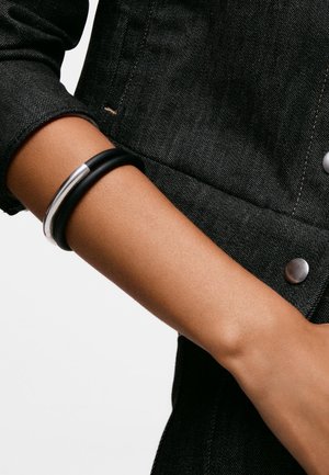 Personne portant un bracelet noir et argenté au poignet, vêtue d'une veste en jean foncée avec des boutons apparents.
