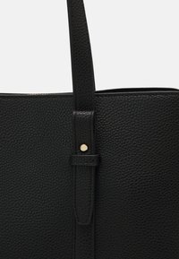 Sac fourre-tout noir en cuir grainé, présentant une sangle lisse et des éléments en métal doré pour la fermeture. Extérieur texturé avec des coutures renforcées.