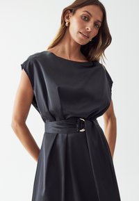 Black satin dress features short sleeves, a wide neckline, and a belted waist with a gold buckle accent. Smooth texture and elegant silhouette.