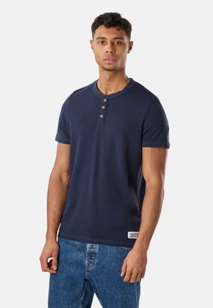 Ung mand iført en marineblå kortærmet henley-shirt med tre knapper, kombineret med blå denimjeans, stående foran en ensfarvet baggrund.