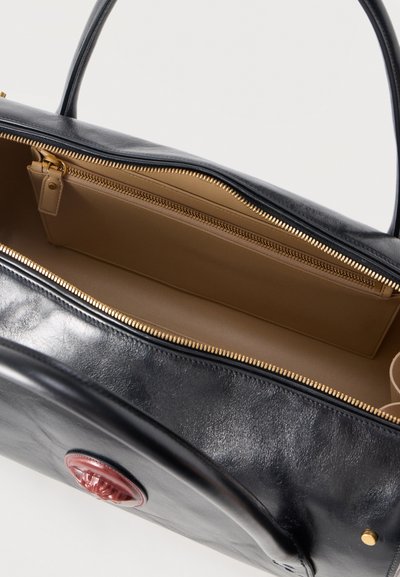 Geantă de mână din piele neagră cu fermoar auriu, interior bej, mânere rotunde și un emblema circulară roșie pe față.