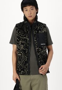Black fleece vest with a paisley pattern, featuring a zip closure and a left chest pocket in solid black fabric. Worn over a gray shirt.