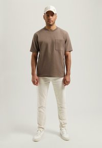 Hnědé tričko s krátkým rukávem a kapsou na prsou, kombinované se světlými slim-fit džíny a bílými teniskami, doplněné bílou čepicí.