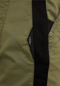 Tissu vert olive à texture lisse, avec un panneau latéral noir, un bouton argenté et une étiquette de logo visible dans la couture.